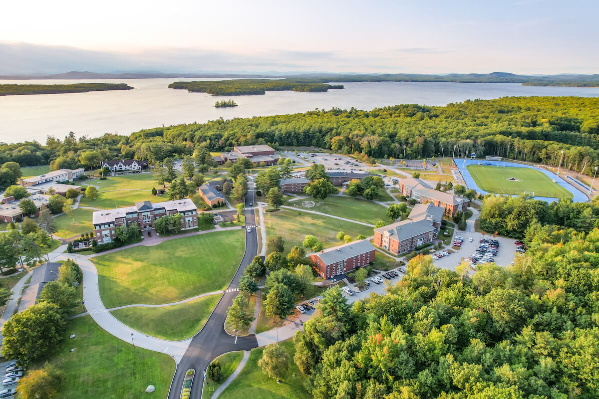 Aerial view of Saint Joseph’s College campus in Maine, showcasing various buildings and sports facilities surrounded by lush greenery, adjacent to a large body of water during daytime. Saint Joseph's College of Maine
