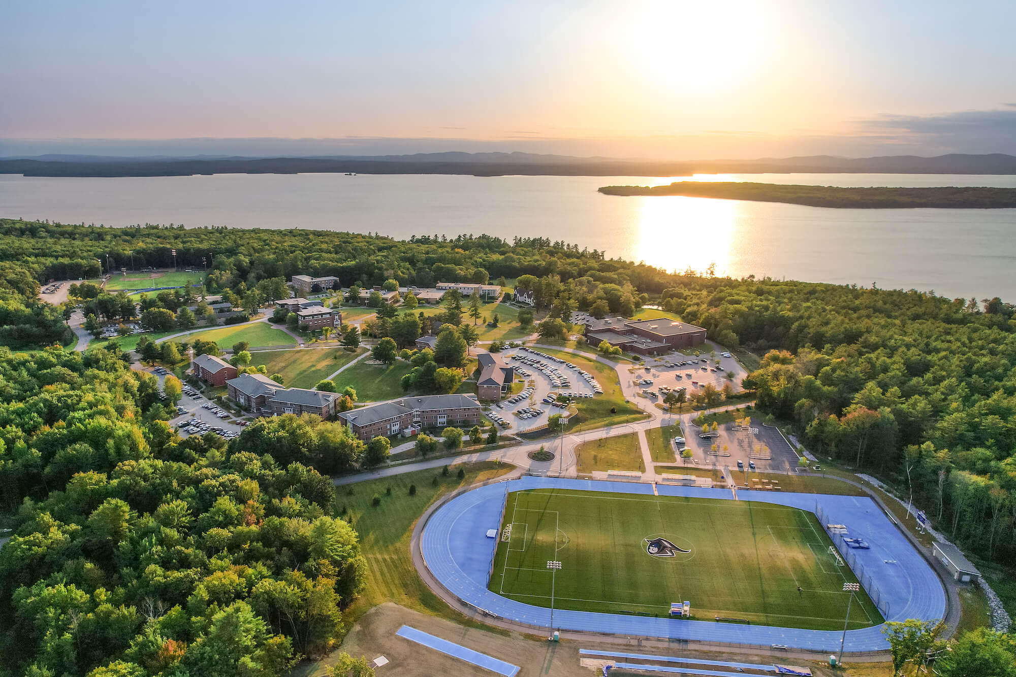Aerial view of a campus with buildings, a running track, and a sports field for aspiring soccer players and Soccer clinics, surrounded by trees near a large body of water at sunset. Saint Joseph's College of Maine