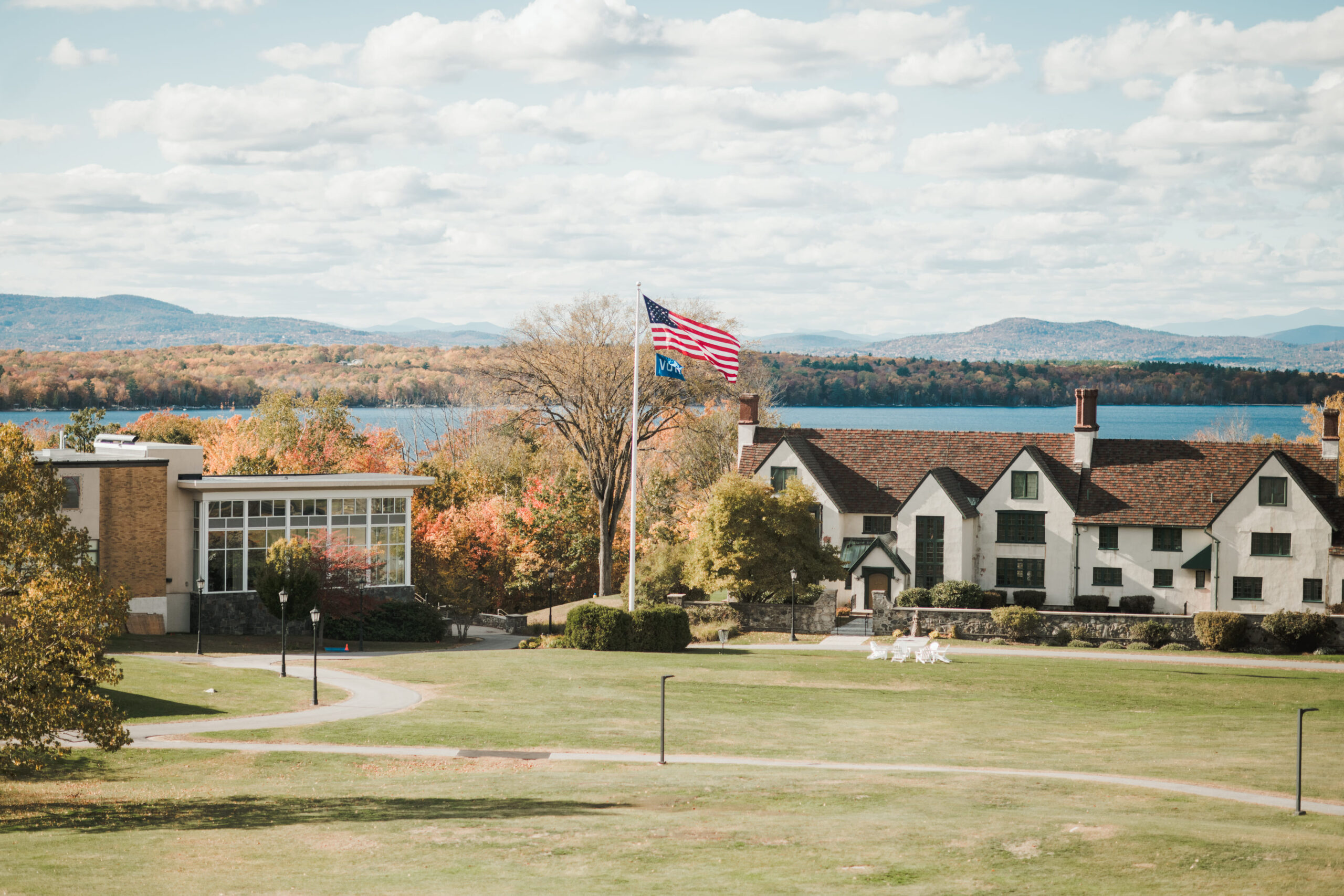 A large American flag flies in front of a Saint Joseph's College of Maine campus building, with a lake, autumn-colored trees, and mountains in the background—a scenic setting for higher education opportunities. Saint Joseph's College of Maine