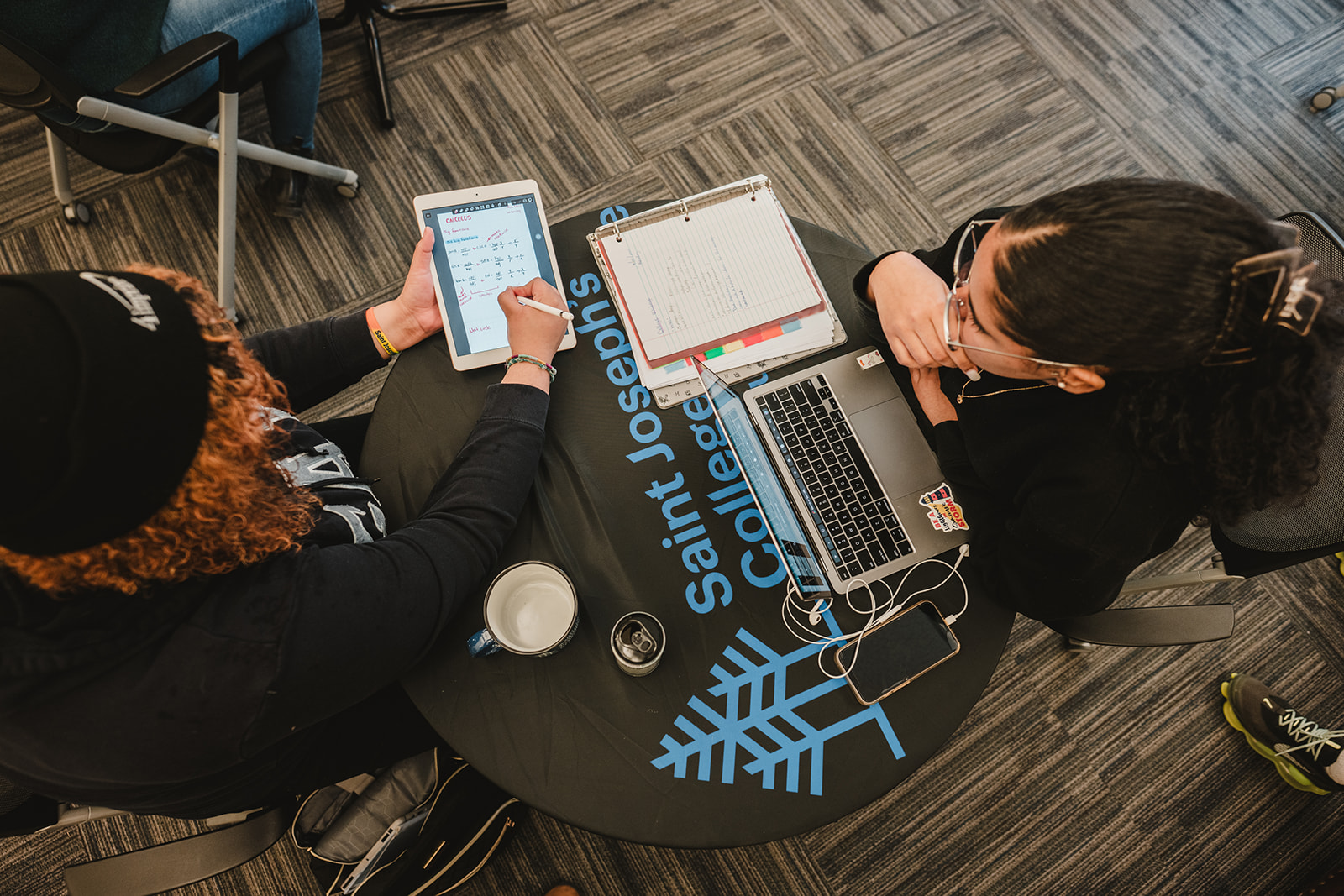 Two people sit at a round table with "Saint Joseph's College" written on it. One holds a tablet and stylus, the other a laptop and notebook. A coffee cup and earphones are also visible on the table, perhaps celebrating making the Dean's List for fall 2023. Saint Joseph's College of Maine