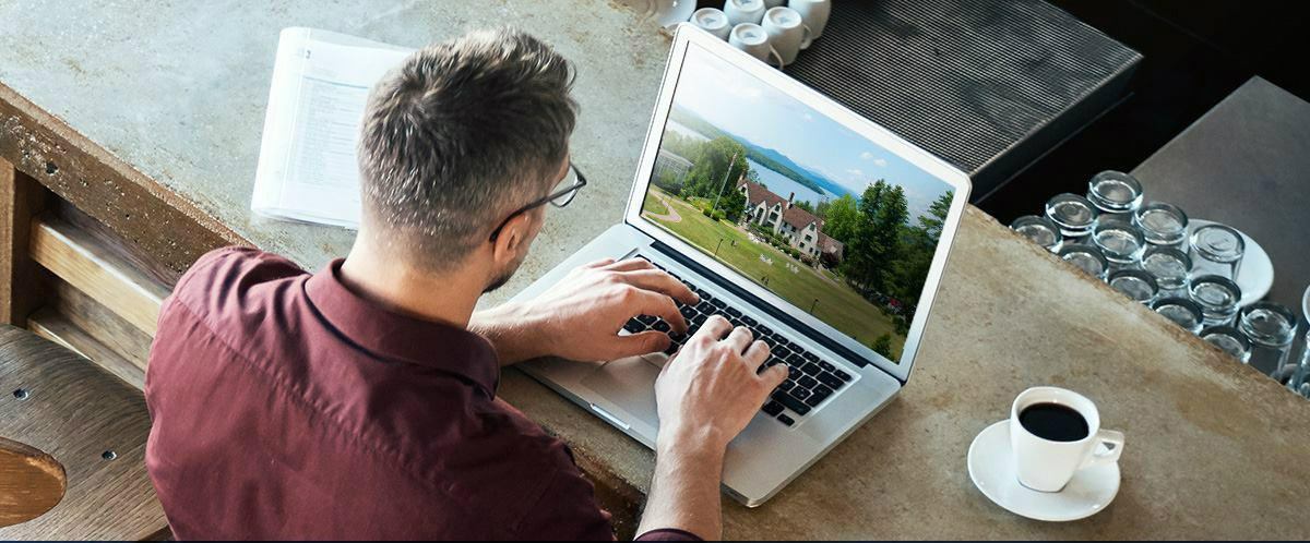 A person with glasses is working on a laptop at a countertop. Next to the laptop, there is a cup of coffee and a book bearing the logo of Saint Joseph's College in Maine. Saint Joseph's College of Maine