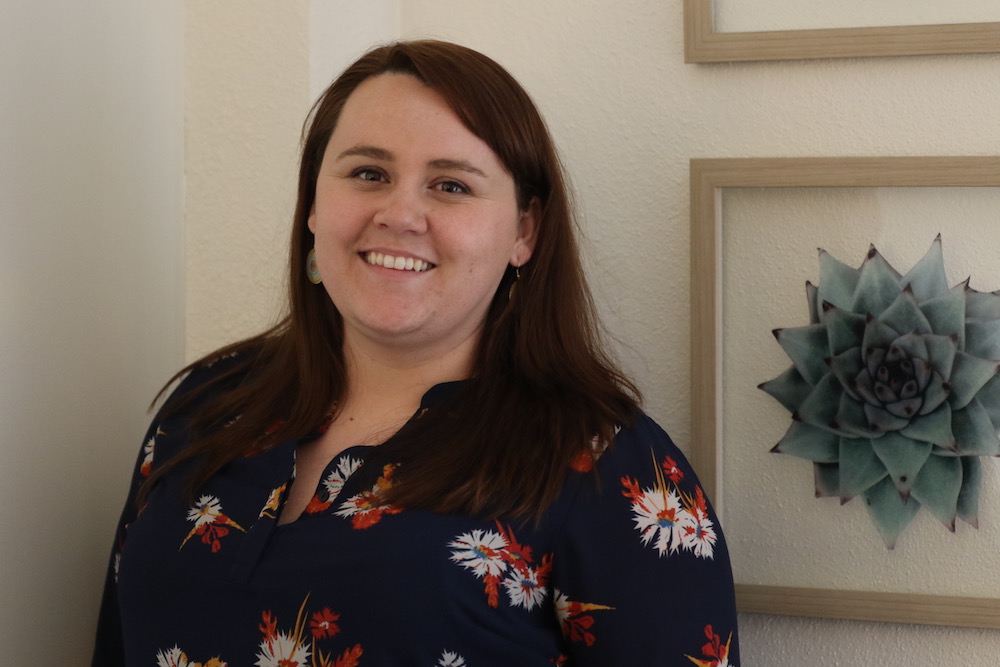 A person with long brown hair, wearing a dark floral top, is smiling in front of a white wall with framed succulent artwork. Their commitment to violence prevention and involvement with the U.S. Department of Justice's grant funds highlight their dedication to creating safer communities. Saint Joseph's College of Maine
