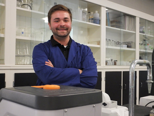 A person wearing a blue lab coat, identified as Raymond Maine, stands with crossed arms in front of laboratory equipment and shelves containing glassware, likely studying infection rates caused by deer ticks. Saint Joseph's College of Maine