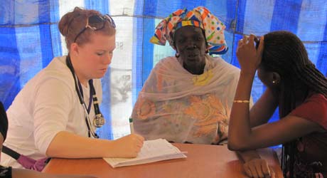 A healthcare worker talks to two women under a blue tent. One woman, dressed in colorful traditional attire from Senegal, listens attentively while the other, possibly student Afar with long braided hair, rests her chin on her hand. Saint Joseph's College of Maine