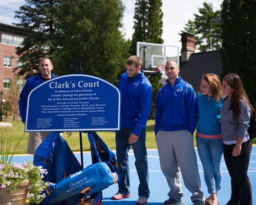 A group of students, some in blue jackets, stand around a blue sign that reads "Clark's Court," as they celebrate on an outdoor basketball court. Saint Joseph's College of Maine