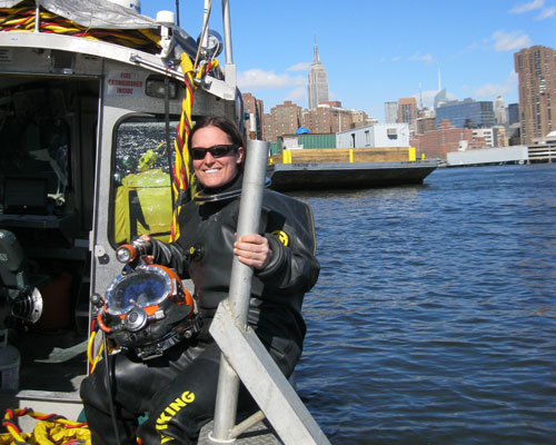 A person wearing a diving suit and sunglasses is happily sitting on the edge of a boat near an urban waterfront, holding diving equipment with a city skyline in the background. Saint Joseph's College of Maine