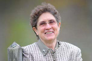 A person wearing glasses and a light-colored, textured jacket is smiling with gratitude while leaning on a wooden post outdoors. Saint Joseph's College of Maine