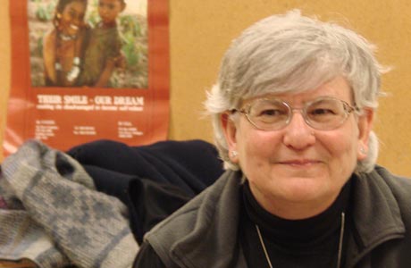 A person with gray hair and glasses sits in front of a poster and a brown backdrop, wearing a black top and a gray vest, reflecting the dedication to the Hispanic community that is evident in this distinguished alumna's work. Saint Joseph's College of Maine