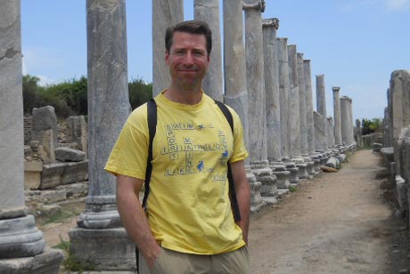 A man in a yellow T-shirt and khaki shorts stands between two rows of ancient stone columns on a dirt path, with a backpack strap visible over his shoulders. As part of The Academy of Travel, he's discovering history firsthand beneath a clear blue sky. Saint Joseph's College of Maine