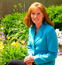 A woman with red hair wearing a blue blazer, Tamera Edison, sits in front of a garden adorned with colorful flowers and lush green plants. An alumni of her university, she reflects on her graduation day amidst the vibrant backdrop. Saint Joseph's College of Maine