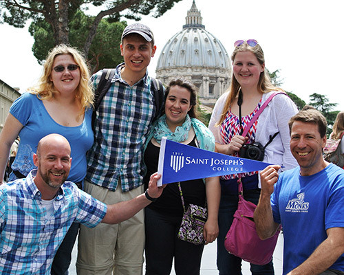 A group of six people are smiling and holding up a "Saint Joseph's College" pennant in an outdoor setting with a domed building in the background, celebrating their Rome Experience. Saint Joseph's College of Maine