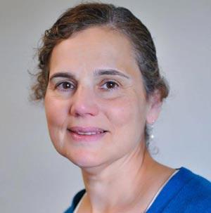 A woman with short, curly brown hair, wearing a blue top, smiles while looking at the camera against a plain background. As an Online Theology Director for her college's certificate programs, she exudes confidence and warmth. Saint Joseph's College of Maine