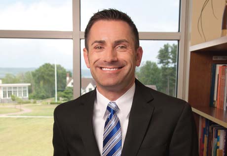 A man in a suit and striped tie is smiling, standing in front of a window with a scenic outdoor view. There is a bookshelf on the right side of the image, adorned with various books. You might recognize him as Vincent J. Kloskowski III, class of '95. Saint Joseph's College of Maine