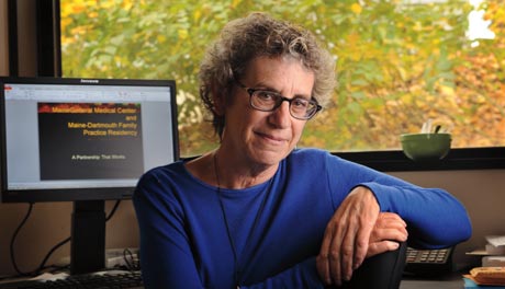 Nancy Weingarten, a person with curly gray hair and glasses, sits at a desk in an office. Behind her is a computer displaying text and a large window with a view of trees, suggesting her residency at MSHA is both peaceful and productive. Saint Joseph's College of Maine