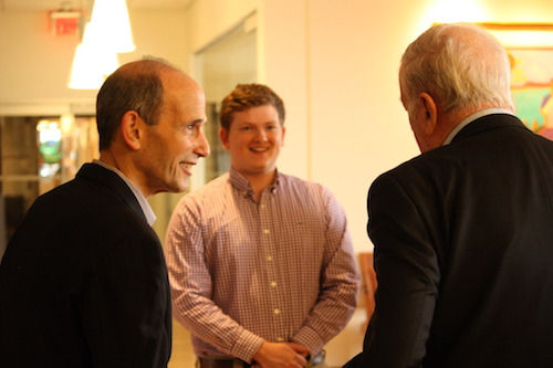 Three men engage in conversation indoors; one older man with a bald head, identified as a Waldron Scholar, is in profile. Another young man in a checkered shirt, a Political Science Major, stands smiling in the background, while the back of a third man's head is visible. Saint Joseph's College of Maine