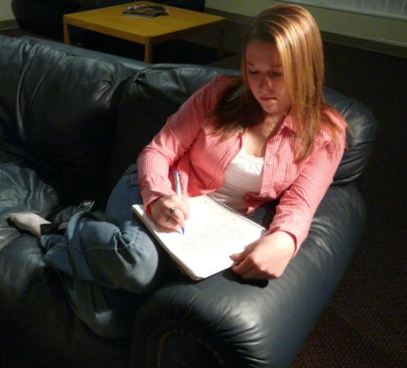 A person with long hair and a red shirt is sitting on a black leather couch, writing a letter in a notebook placed on their lap. Saint Joseph's College of Maine