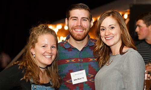 Three people are standing together and smiling at an indoor alumni event. The person in the middle has a name tag. There are string lights in the background, adding a warm glow to the atmosphere as they gather to raise funds for scholarship opportunities. Saint Joseph's College of Maine
