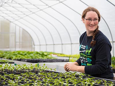 A woman with glasses and a braid tends to seedlings in a greenhouse filled with rows of young plants, preparing for Pearson’s Town Farm's first seedling sale. Saint Joseph's College of Maine