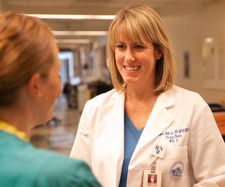 A nurse in a white coat with an RN badge chats with a colleague in green scrubs in a hospital hallway, perhaps discussing the radical notion of extending their lunch break. Saint Joseph's College of Maine