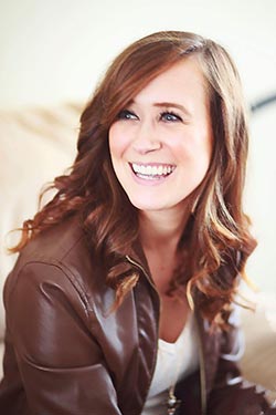 A woman with wavy brown hair is smiling and looking to the side. She is wearing a brown leather jacket over a white shirt, possibly reflecting on her recent commencement from an online program. Saint Joseph's College of Maine