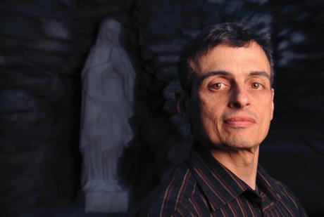 A man in a striped shirt stands in front of a blurred statue with a neutral expression, perhaps searching for a little miracle. The background is dark, and the statue appears to be of a standing figure. Saint Joseph's College of Maine
