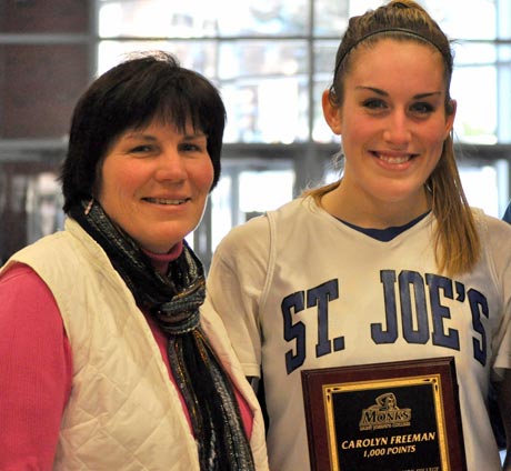 Two women standing side by side, one in a white vest and scarf, the other in a "St. Joe's" sports uniform holding a plaque that reads "Carolyn Freeman, 1,000 Points". Their smiles reflect the pride of achieving such high scoring runs within their supportive family. Saint Joseph's College of Maine