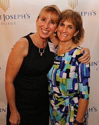 Two women are smiling and standing together in front of a Saint Joseph's College banner. One, Catherine Renquin Ryder, is wearing a black dress, while the other is in a colorful geometric patterned dress. Their joy likely stems from celebrating the Professional Excellence Award at Saint Joseph’s College. Saint Joseph's College of Maine