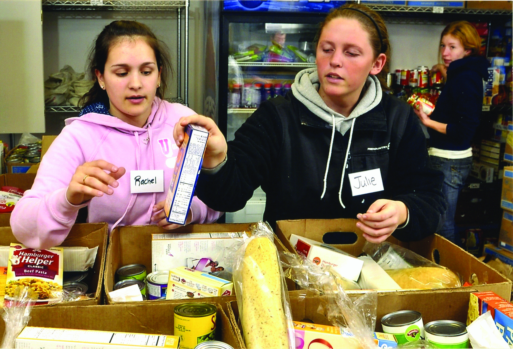 Two women, wearing name tags reading "Rachel" and "Julie", sort food items in boxes at a food bank. Reflections of their commitment glimmer as they work diligently. Another person in the background also handles food items, ensuring no cupboard is ever empty. Saint Joseph's College of Maine