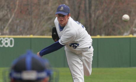 A power pitcher in a white and blue uniform throws a pitch towards the batter on a grass field, with the catcher seen in the foreground. Saint Joseph's College of Maine