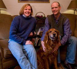 A woman and a man sit on a couch with their golden retriever, Cassie, nestled between them, all smiling. Saint Joseph's College of Maine