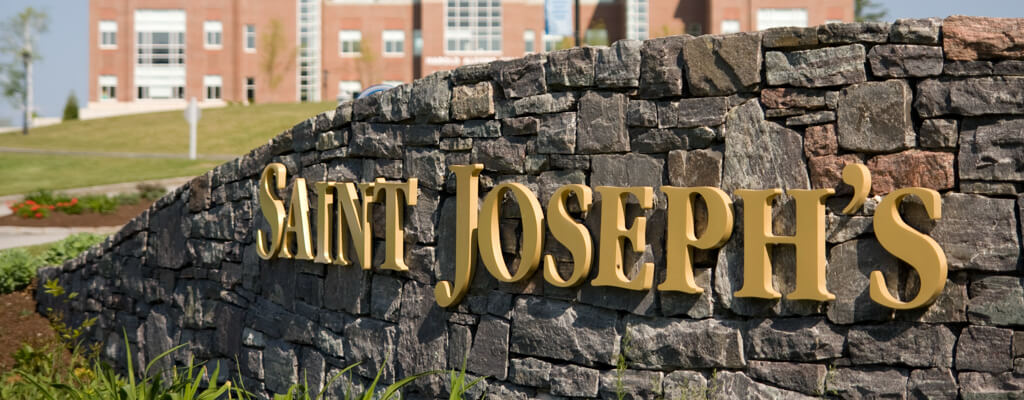 Golden "Saint Joseph's" letters are mounted on a stone wall in the foreground, with a brick building visible in the blurred background. This setting stands as a testament to the achievements of the SJC Science Scholars Program, supported by National Science Foundation funding. Saint Joseph's College of Maine