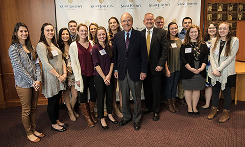 A group of people, consisting of students and adults in professional attire, stand together in front of a Saint Joseph's College backdrop, celebrating an extraordinary opportunity as Mitchell Scholars with Senator George Mitchell. Saint Joseph's College of Maine