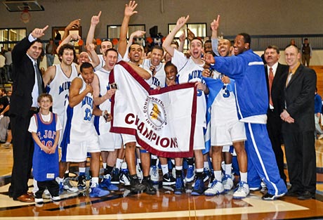 A men's basketball team celebrates on the court, holding a "Champion" banner. Coaches, staff, and a child in a jersey stand with them as they savor their championship win. Some players and coaches raise their hands in victory, capturing the hearts of everyone present. Saint Joseph's College of Maine