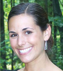 A double alumni with dark hair tied back, wearing earrings and a black top, smiles at the camera with a forested area in the background. Saint Joseph's College of Maine