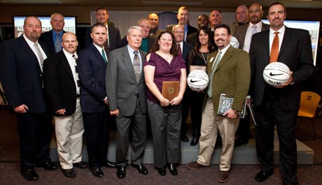 A group of people, dressed in formal and business attire, are standing together in a room. Two individuals in the front row are holding basketballs, likely honored for their athletics achievements with a Hall of Fame induction in 2012. Saint Joseph's College of Maine