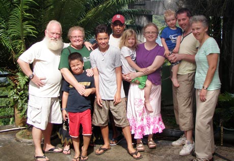 Group photo of ten people, including children and adults, standing outdoors in front of lush greenery. Everyone is smiling, several are holding or hugging each other, and the alumna who traveled the farthest away for this gathering is positioned proudly at the center. Saint Joseph's College of Maine