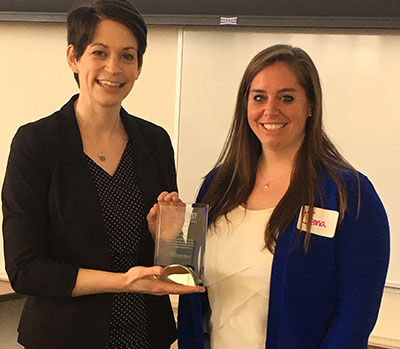 Two women are standing indoors. One, Jenna Chase, holds an award while the other stands beside her, smiling. Both are dressed in business attire, representing the Breathe Easy Coalition during a college event. Saint Joseph's College of Maine