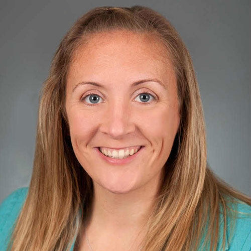 A woman with long blonde hair, wearing a teal top, smiles at the camera against a gray background, capturing one of those light bulb moments. Saint Joseph's College of Maine