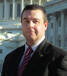 A man in a black suit and red tie, whose confident demeanor catches the eye, stands in front of a large classical building with columns on a sunny day. Saint Joseph's College of Maine