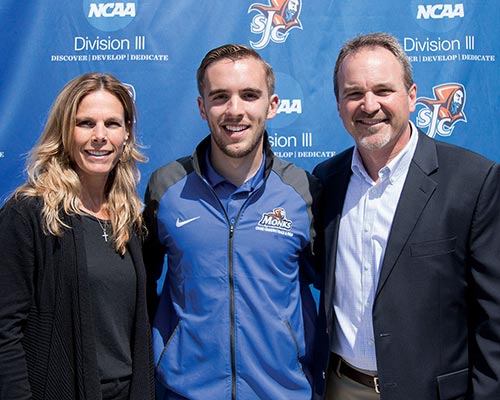 Three people standing in front of a blue NCAA Division III backdrop: a woman on the left, a man in athletic wear in the center, and a man in business attire on the right, embodying the Big Tent philosophy that unites diverse talents and backgrounds. Saint Joseph's College of Maine