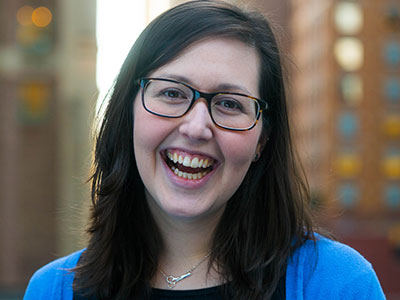 Kerry Weber, a Catholic journalist with long dark hair and glasses, is seen smiling in a blue cardigan and necklace, standing in front of a blurred urban background. Saint Joseph's College of Maine