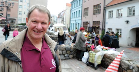 Mark Friedman, a smiling man in a maroon shirt and beige jacket, stands in a cobblestone plaza with market stalls and people bustling in the background. Saint Joseph's College of Maine