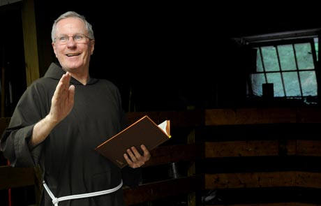 A man in a brown robe with a white cord, likely a chaplain from the Mercy Center, holds an open book and gestures with his hand, standing in a dimly lit room with a window in the background. Saint Joseph's College of Maine