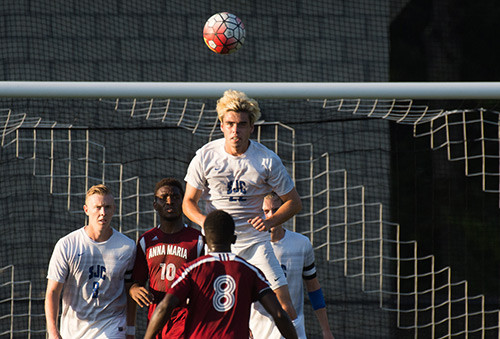 A soccer player in a white jersey from the 8th regionally ranked men's soccer team heads the ball near the goal while three other players, two in white and one in red, observe. Saint Joseph's College of Maine