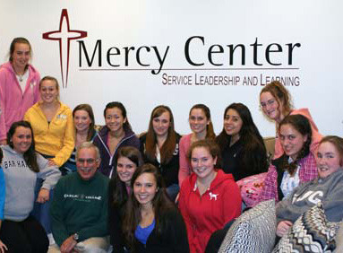 A group of 15 people pose for a photo in front of a wall displaying the Mercy Center logo and the tagline "Service, Leadership, and Learning," embodying the spirit of faith and service. Saint Joseph's College of Maine