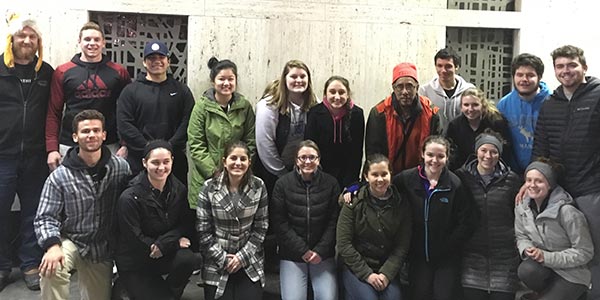 A group of 18 students, wearing winter clothing, pose together indoors in front of a plain background, preparing for the Midnight Run. Saint Joseph's College of Maine