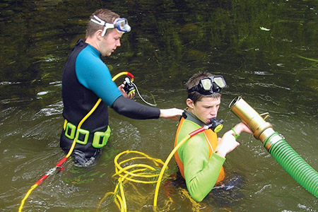 Two divers in wetsuits and goggles stand in shallow water; one holds a hose, while the other adjusts equipment on his chest as part of a $500 college consortium project combating invasive plant species. Saint Joseph's College of Maine