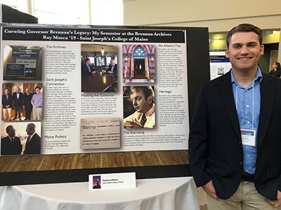 A person stands beside a display titled "Curating Governor Brennan’s Legacy: My Semester at the Brennan Archives," featuring various photos and text panels. As part of the Statewide Education Consortium, the person holds a certificate while smiling at the camera, celebrating their achievement. Saint Joseph's College of Maine