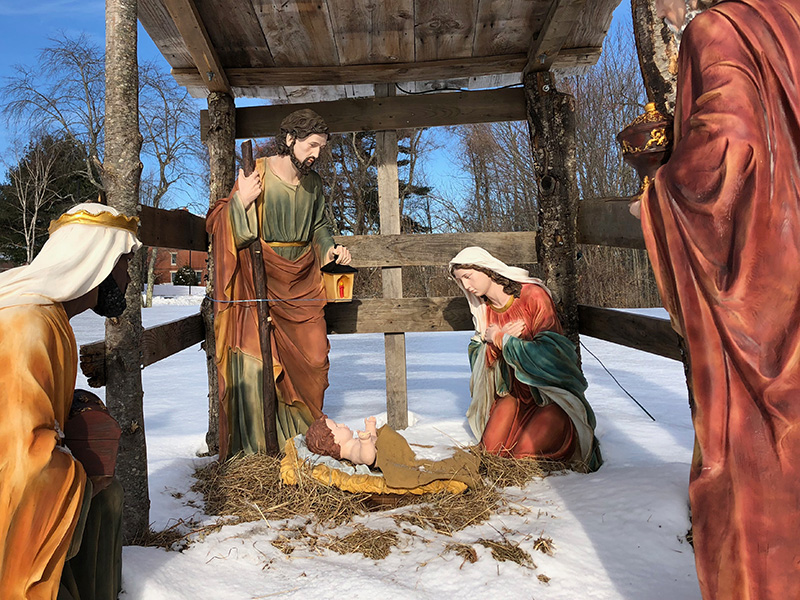 Outdoor Nativity scene in the snow, featuring statues of Mary, Joseph, baby Jesus in a manger, and two other figures kneeling. Saint Joseph's College of Maine