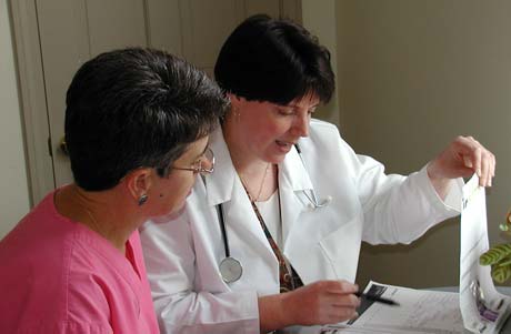 A doctor in a white coat shows documents to a person in a pink shirt while discussing medical information at a table, also mentioning the benefits of online graduate programs for continuing education. Saint Joseph's College of Maine